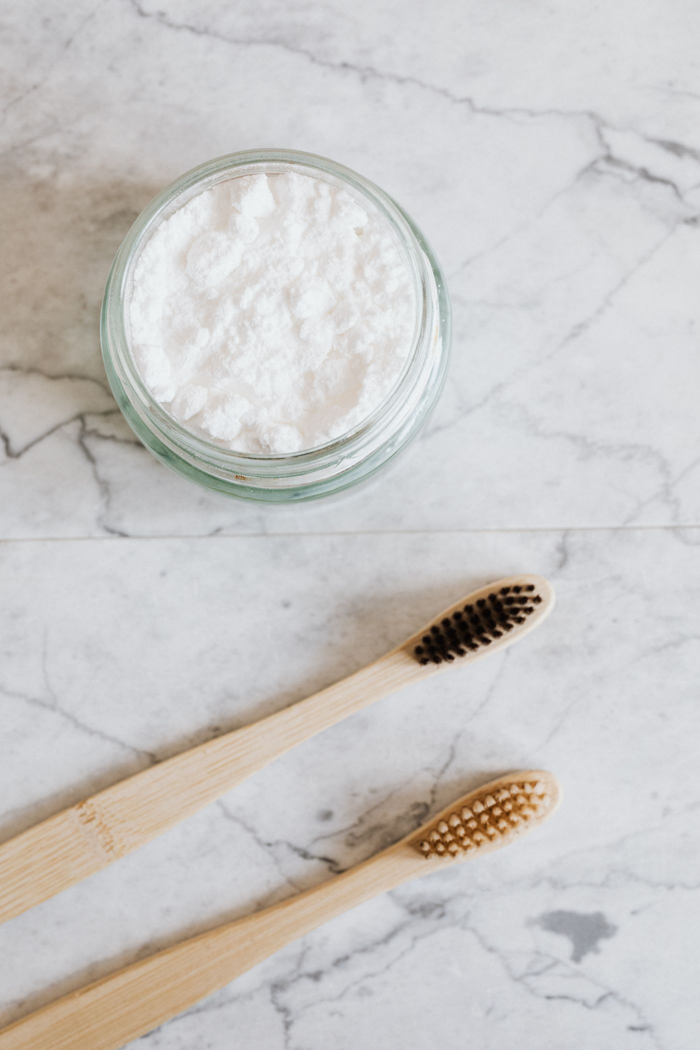 An open glass jar of white tooth powder beside a toothbrush on marble